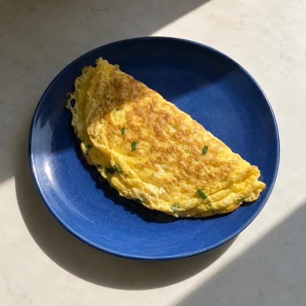 A folded golden omelet with green chive pieces sits on a cobalt-blue ceramic plate on a pale stone-like table.