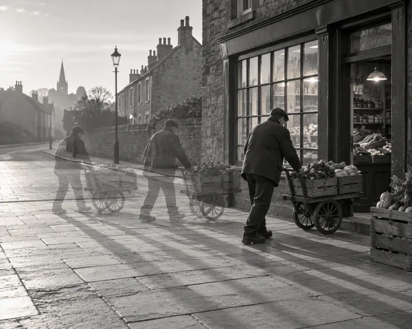 A black-and-white street scene shows a produce vendor in three successive positions pushing a wooden vegetable cart toward a lit neighbourhood shop; the two…