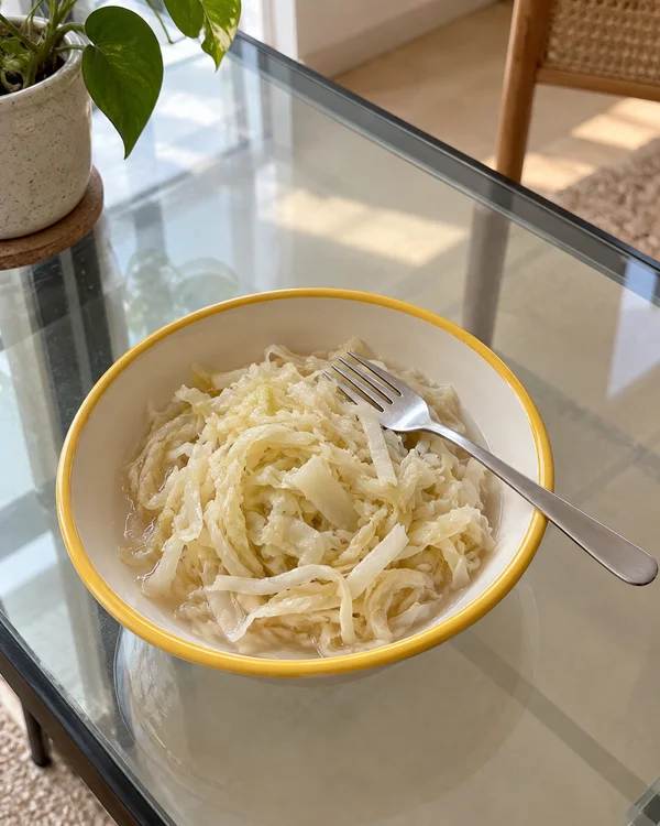 A cream bowl with a yellow rim holds pale shredded cabbage on a glass table, with a stainless-steel fork laid across it.