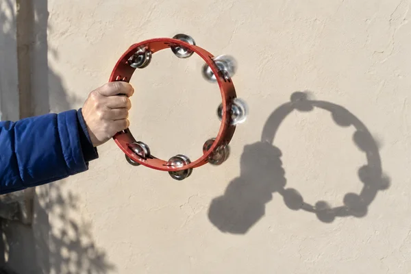 A hand in a cobalt-blue winter jacket holds a red tambourine in front of a sunlit cream plaster wall, with its circular shadow falling to the right.