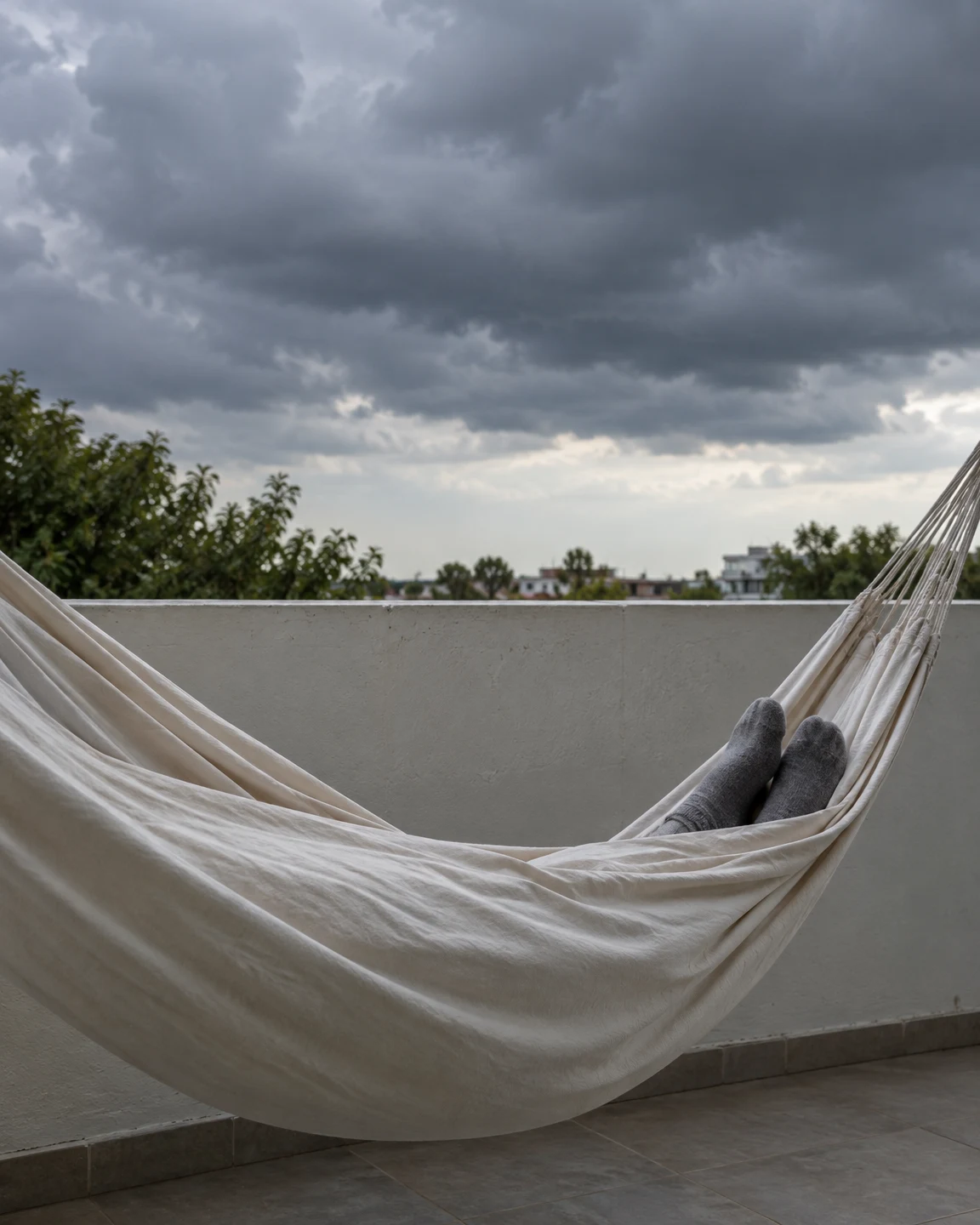 A cream canvas hammock hangs across a tiled home balcony beneath dark storm clouds, with two grey-socked feet resting near its right edge.