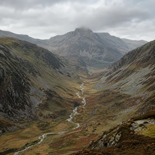 A narrow silver stream winds through a long mountain valley between steep rocky slopes, leading toward a rugged peak partly covered by low grey cloud.