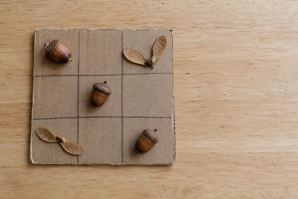 An overhead view of a handmade tic-tac-toe board drawn on brown cardboard, with three acorns and two paired maple seeds on a pale wooden table and open…