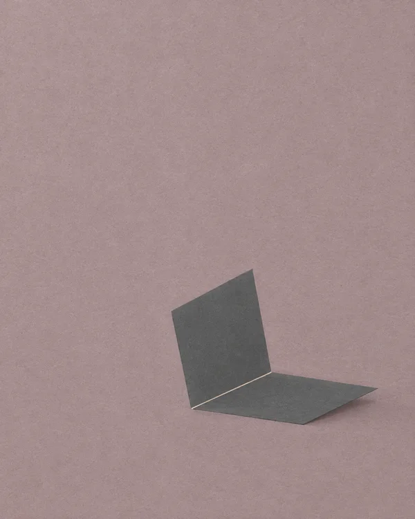 Two dark graphite angular planes meet along a thin pale seam near the lower right-centre of a dusty mauve, paper-textured background, with a soft shadow…