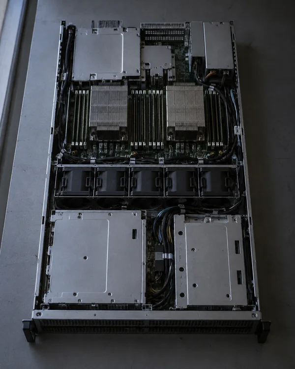 An open rackmount server rests lengthwise on a grey workbench, showing rows of black fans, two broad heat sinks, metal compartments and routed cables under…