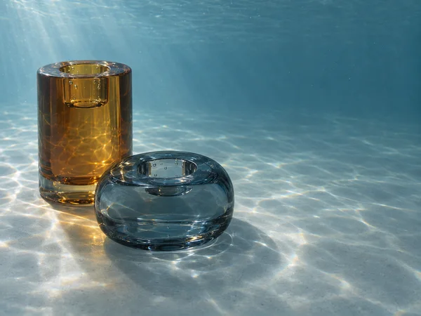 Two solid glass candleholders stand on a submerged blue-grey floor: a tall amber cylinder behind a low smoke-blue rounded holder.
