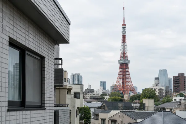 A vermilion-and-white Tokyo Tower rises above a dense mix of residential rooftops and mid-rise buildings beneath a pale overcast sky.