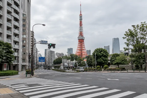 Tokyo Tower rises above an urban intersection, with a broad diagonal zebra crossing in the foreground, light vehicle traffic, roadside buildings, trees, and…
