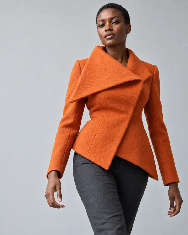 A woman with closely cropped hair wears a vivid orange sculptural jacket with an oversized triangular folded lapel and charcoal trousers against a pale grey…