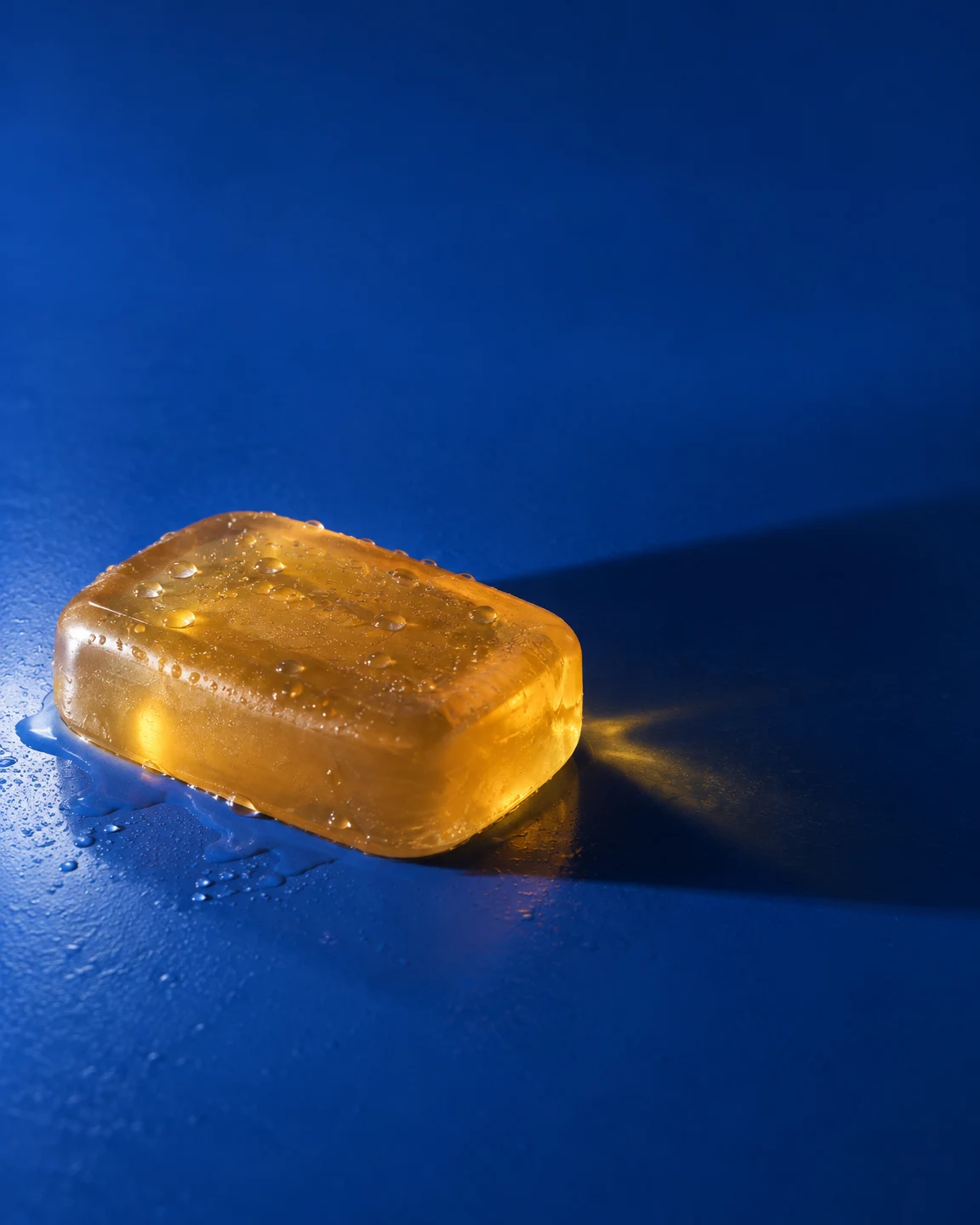 A translucent amber rectangular cleansing bar dotted with water beads rests on a cobalt-blue surface.