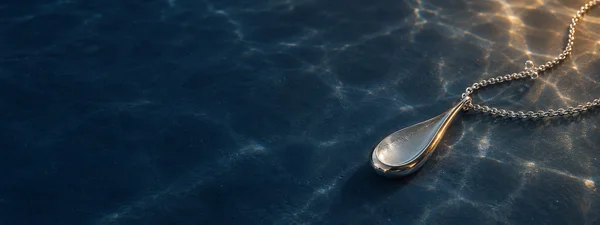 A reflective silver teardrop pendant and fine chain rest on a deep blue underwater surface, with warm highlights, rippling light patterns and a few bubbles…