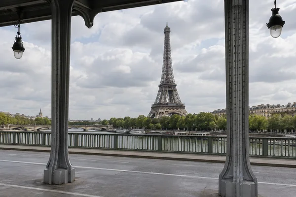 The Eiffel Tower rises above riverside trees and buildings across the Seine, framed by two riveted iron columns beneath a dark bridge deck; a metal railing and…