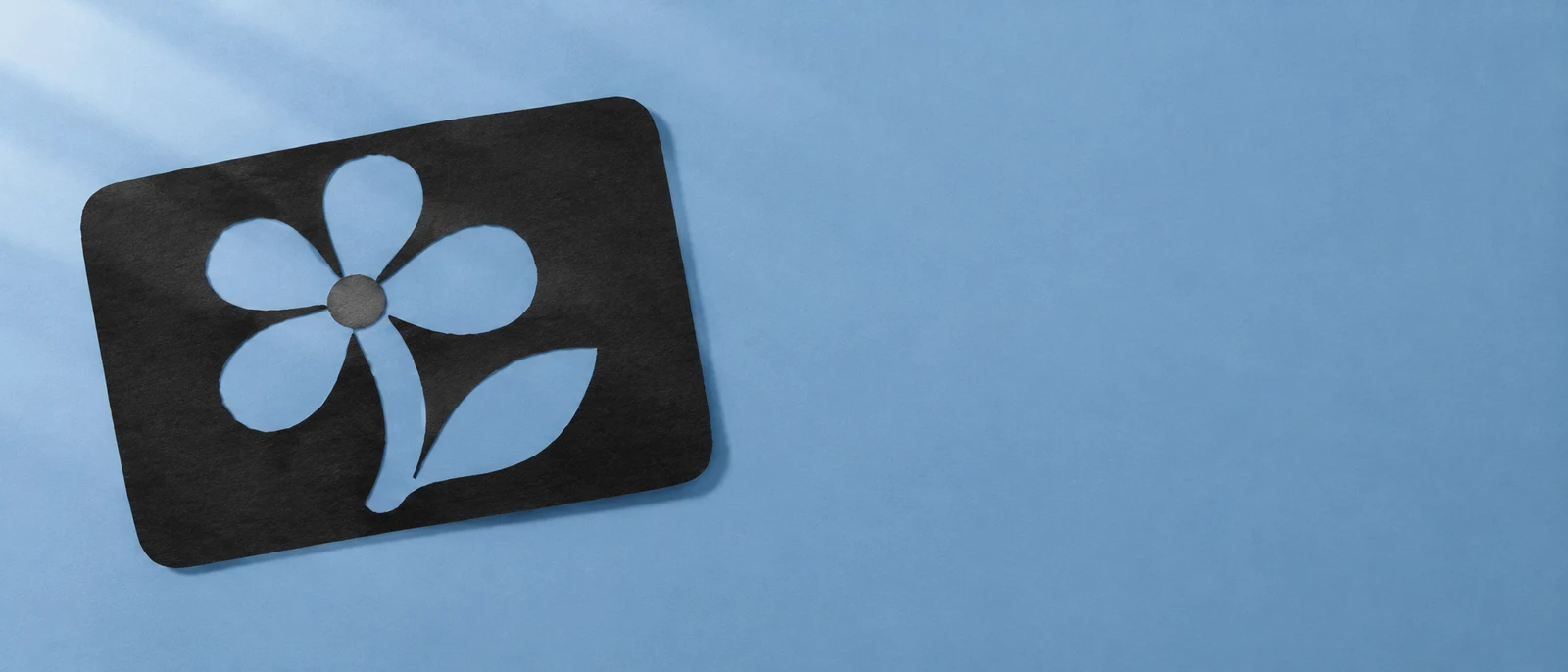 A textured black paper card lies at an angle on a pale blue surface, with a flower-and-leaf shape cut into it and open space to the right.