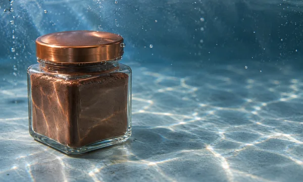A square glass jar of dark-brown powder with a copper-colored lid sits on a pale blue-gray floor underwater, with light patterns and bubbles around it and open…