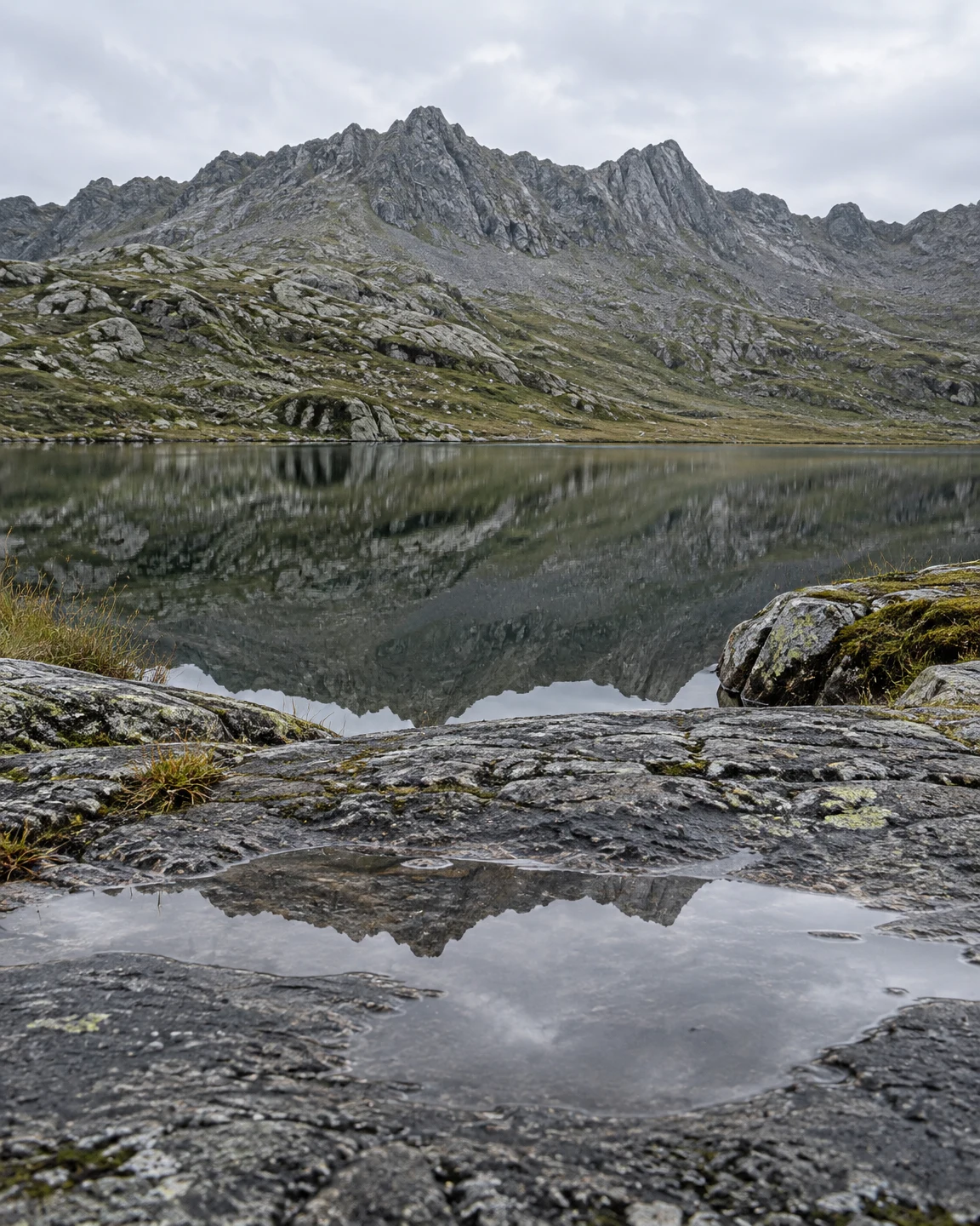 A rugged grey mountain ridge rises above a still tarn, with the ridge reflected in the lake and again in a shallow film of water across a rain-wet foreground…