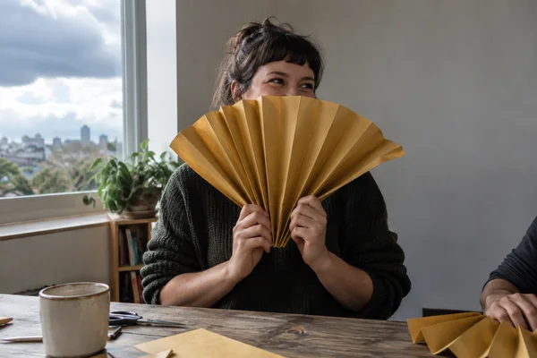 A woman seated at a wooden table holds a large ochre accordion-folded paper fan across her chest and lower face, looking toward a companion whose arm and hand…