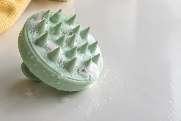 A mint-green silicone scalp brush covered with white shampoo lather lies teeth-up on a wet white tabletop, with water droplets nearby and a yellow textured…