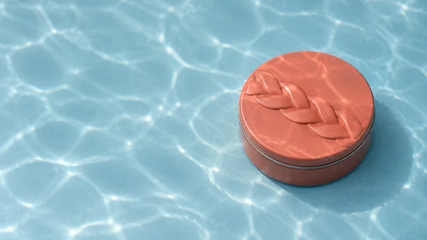 A round coral-colored metal tin with a raised braid motif on its lid sits on a pale blue floor patterned with bright water caustics, with open space to the…