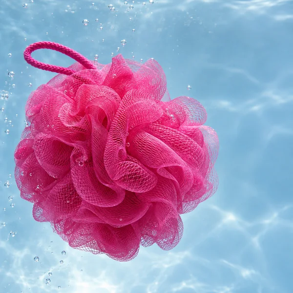 A bright pink mesh shower pouf fills the left side of a pale blue underwater scene, with a long matching loop arcing above it and numerous bubbles around it.