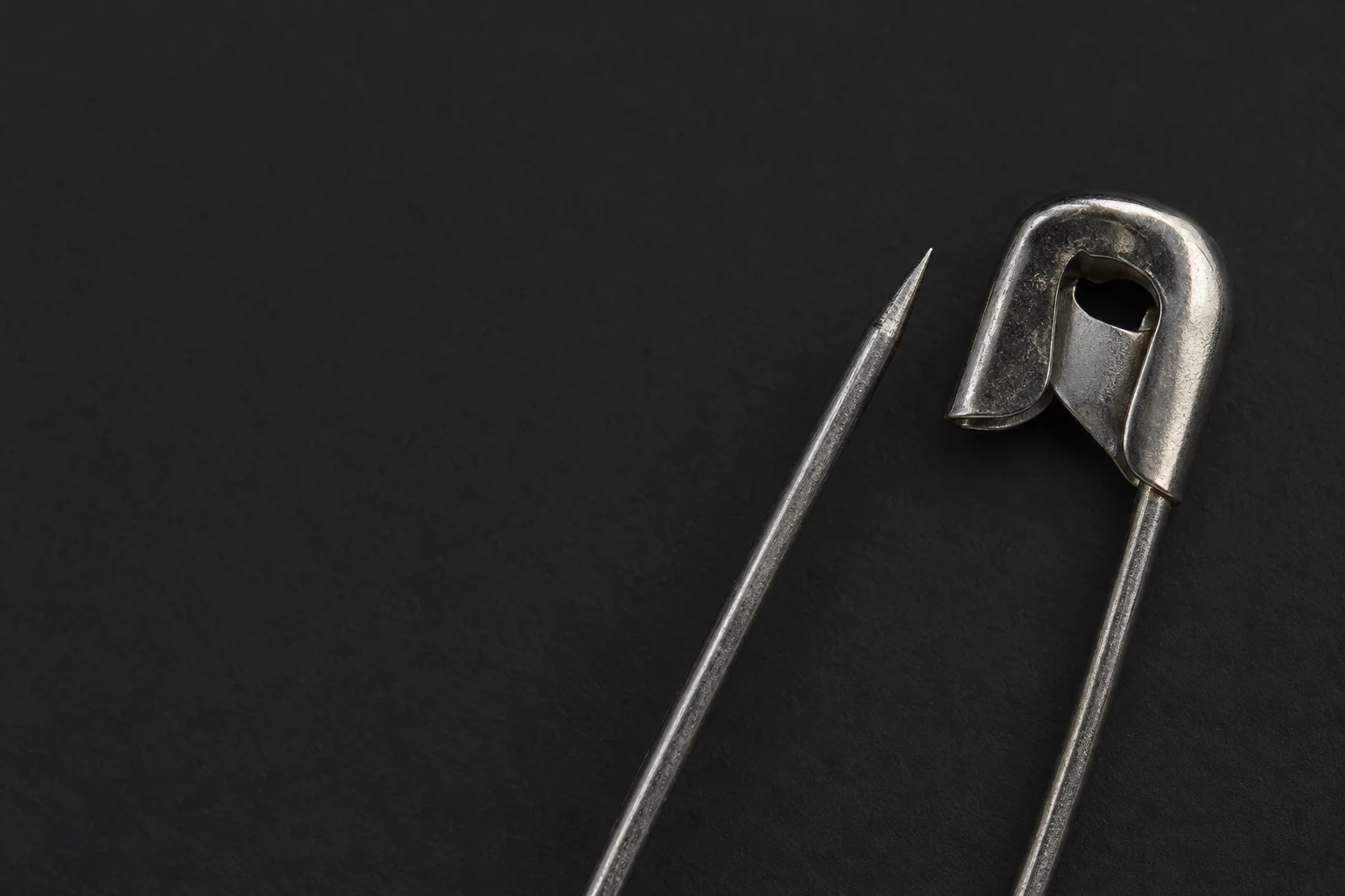 A close-up of an open steel safety pin against a dark charcoal surface, with its pointed end and broad clasp in the upper-right area and the wire extending out…