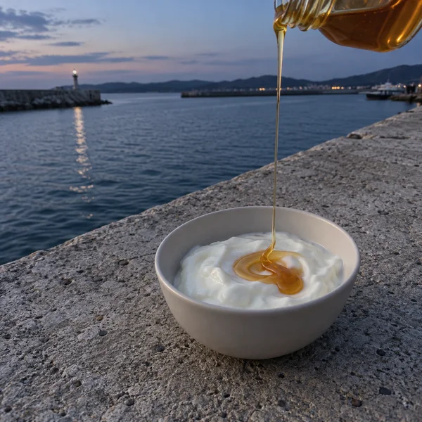 A white bowl of yogurt sits on a rough concrete harbour parapet as amber honey pours from a clear jar cropped at the upper right.