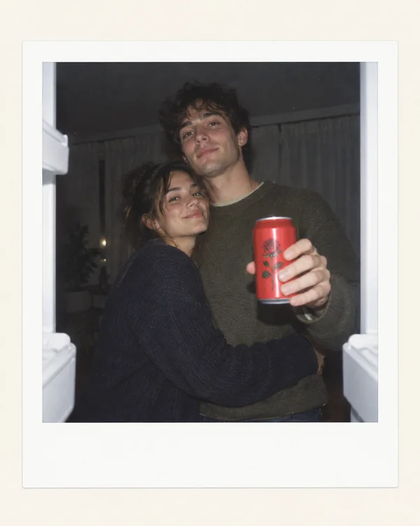 A woman embraces a man around the waist as he holds a red can with a rose illustration toward the camera, seen from inside an open refrigerator.