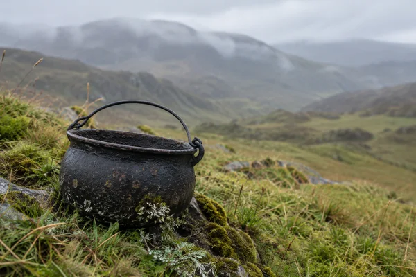 A weathered black cast-iron pot with an arched handle sits among moss, lichen, rocks and tussock grass on a hillside, with softly blurred rolling hills behind…