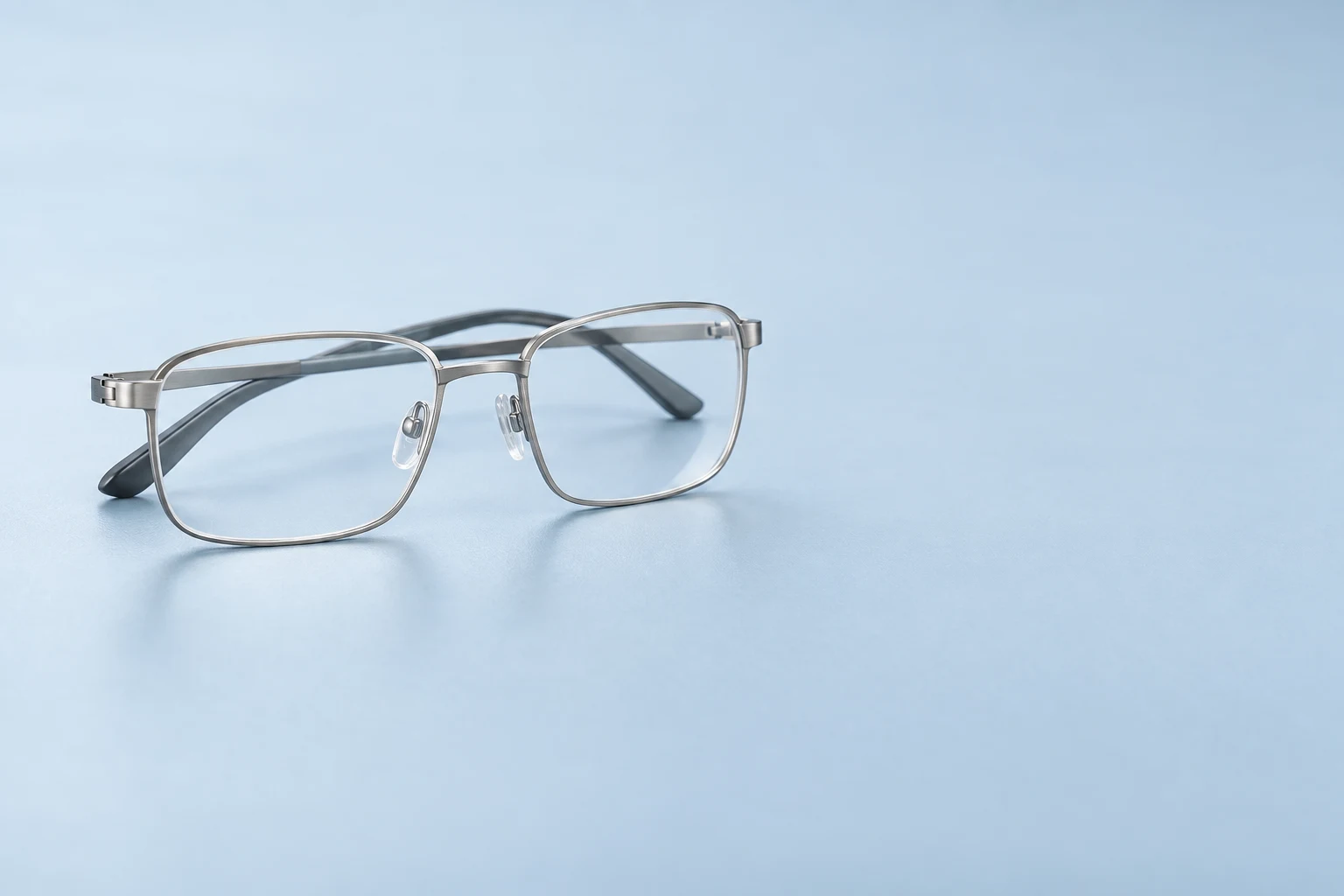 Silver rectangular eyeglasses with clear lenses, transparent nose pads and dark temple tips rest on a pale blue surface, with broad empty space to the right.