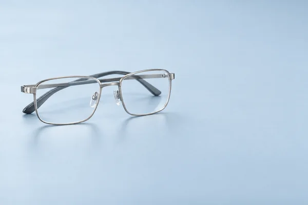 Silver rectangular eyeglasses with clear lenses, transparent nose pads and dark temple tips rest on a pale blue surface, with broad empty space to the right.