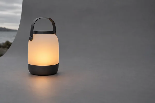 A portable lantern with a dark charcoal base and handle glows warm amber on a slate-grey surface, with a strip of cloudy sky and water visible beyond the…