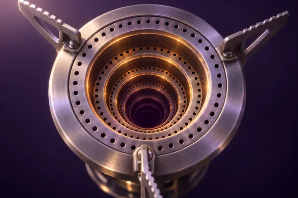 A close-up of a brushed-metal camp-stove burner head with three serrated pot supports and concentric perforated rings receding into an amber-lit, dark plum…