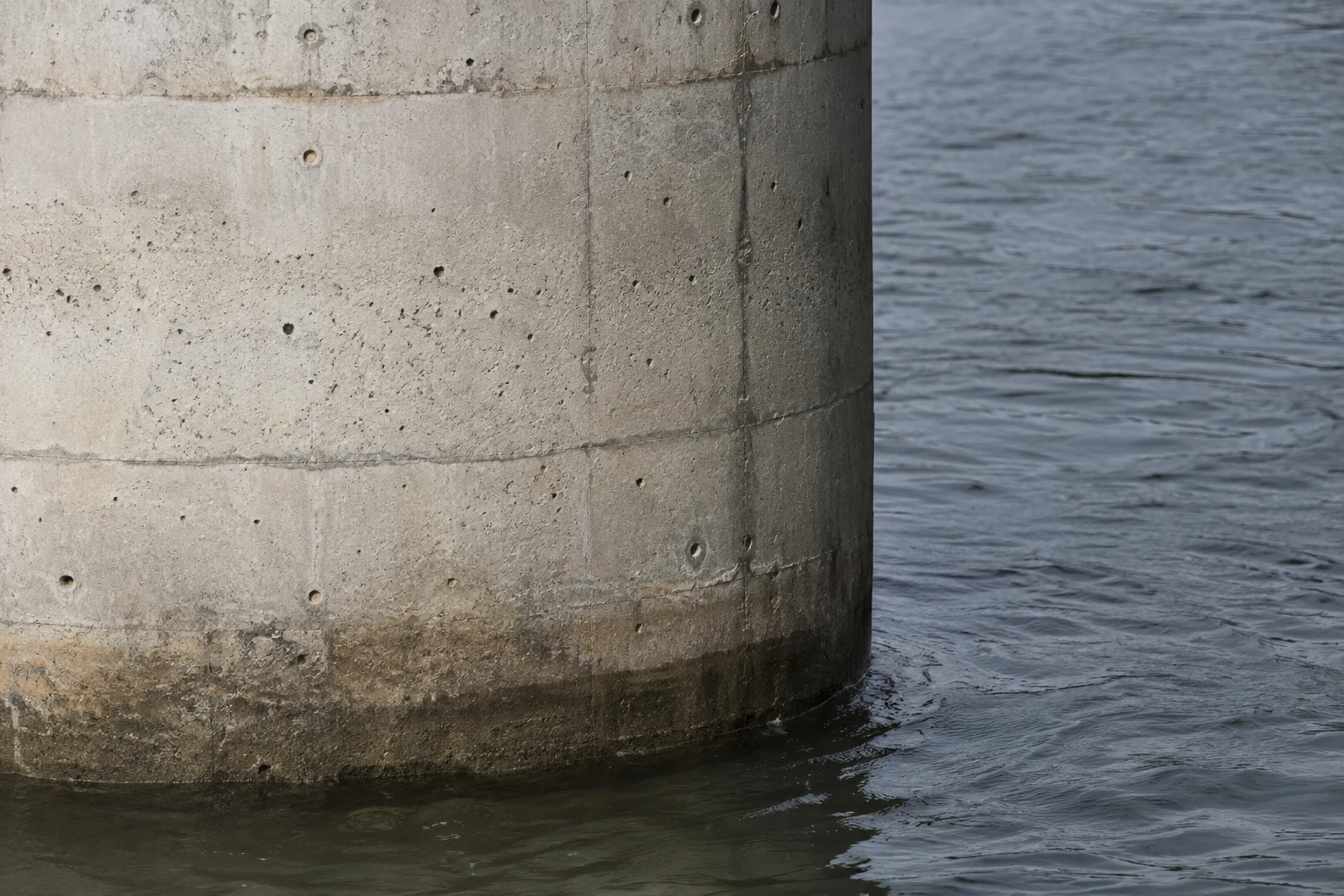 Close view of a pale concrete bridge pier meeting rippling slate-grey water, with horizontal casting seams, small air holes and a dark waterline.