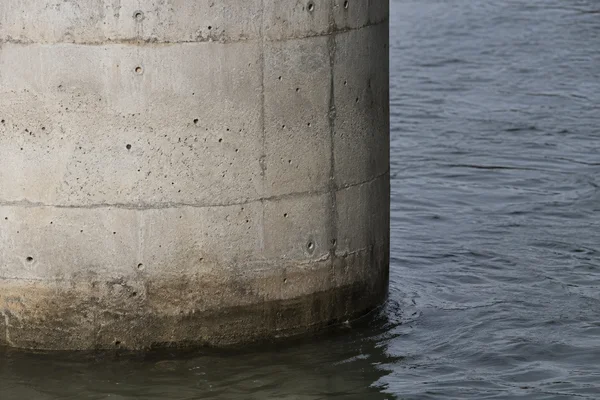 Close view of a pale concrete bridge pier meeting rippling slate-grey water, with horizontal casting seams, small air holes and a dark waterline.