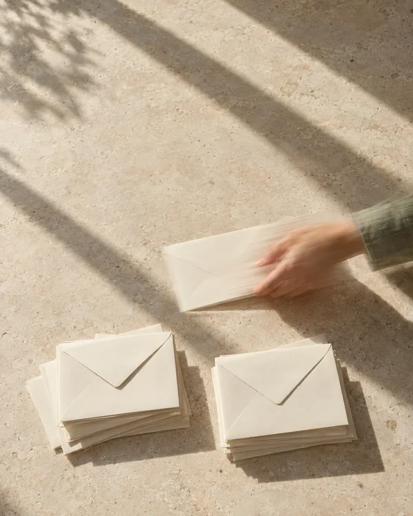A hand in a sage-green cuff slides an ivory envelope across a pale limestone surface toward two small piles of envelopes, with strong diagonal sunlight and…