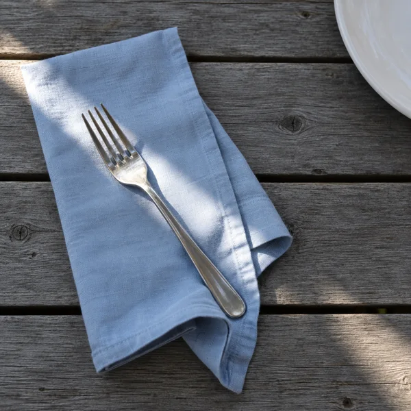 A pale-blue folded cotton napkin and stainless-steel fork lie on weathered grey wooden boards, with part of a white ceramic plate entering from the upper-right…