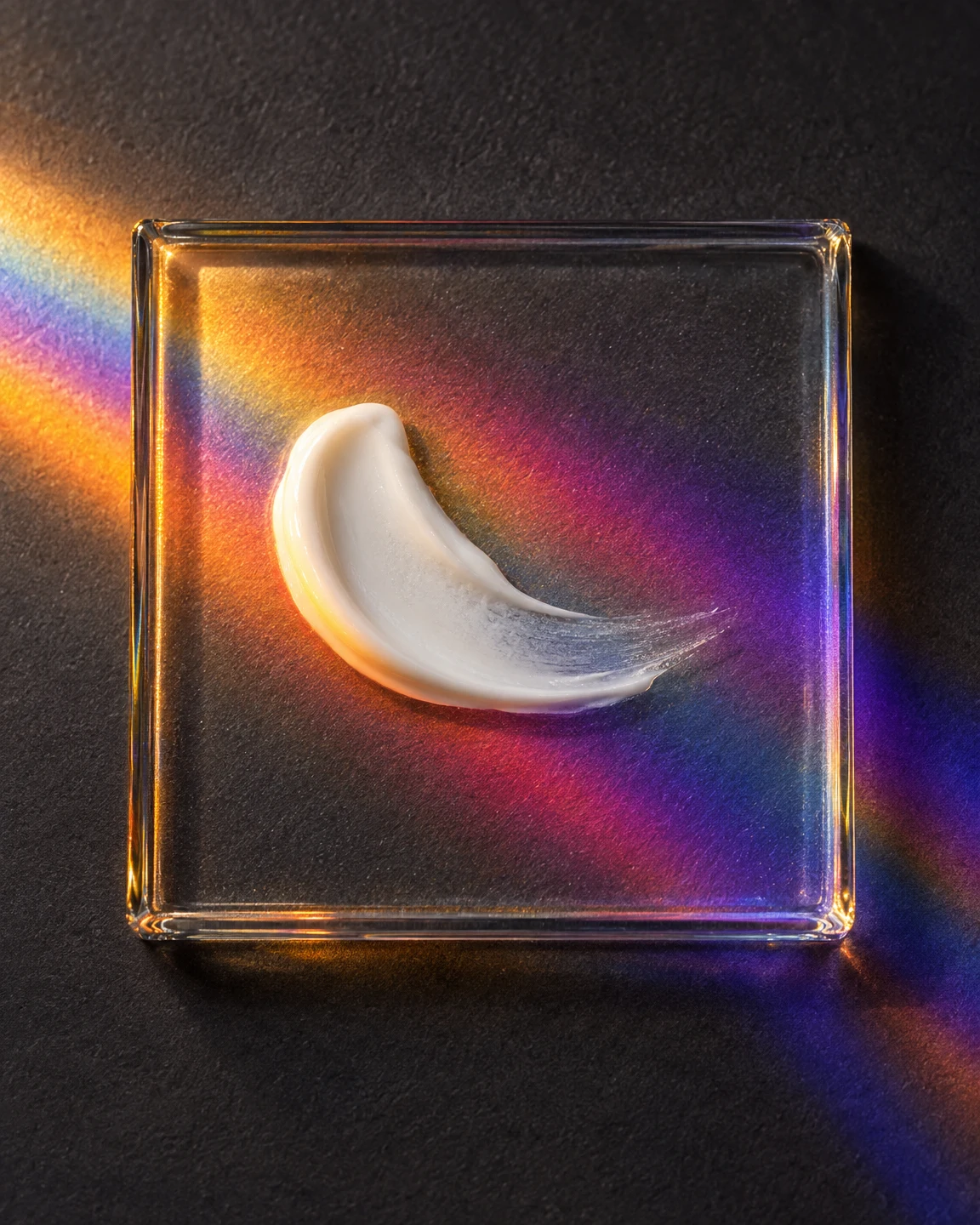 A curved white cream smear rests on a clear square glass tile against a charcoal surface, surrounded by amber, magenta and violet bands of light.