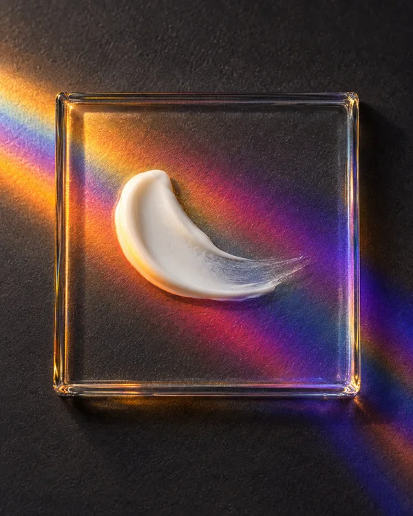 A curved white cream smear rests on a clear square glass tile against a charcoal surface, surrounded by amber, magenta and violet bands of light.