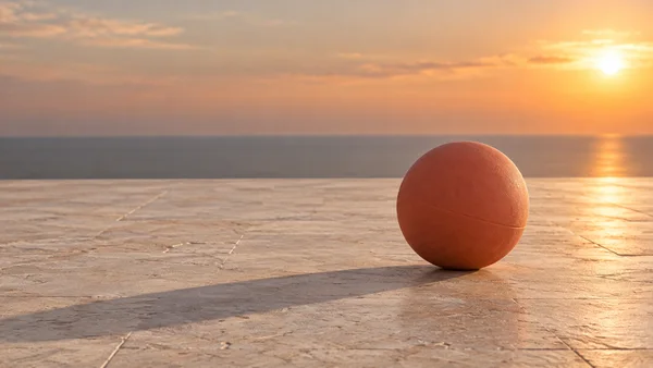A terracotta-colored rubber ball rests on a textured stone terrace at sunset, with a long shadow stretching toward the lower left and water visible beyond the…
