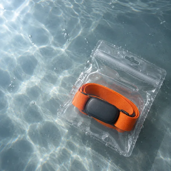 An orange elastic chest strap wrapped around a dark graphite heart-rate sensor sits inside a clear pouch on a blue-grey tank floor underwater, with bright…