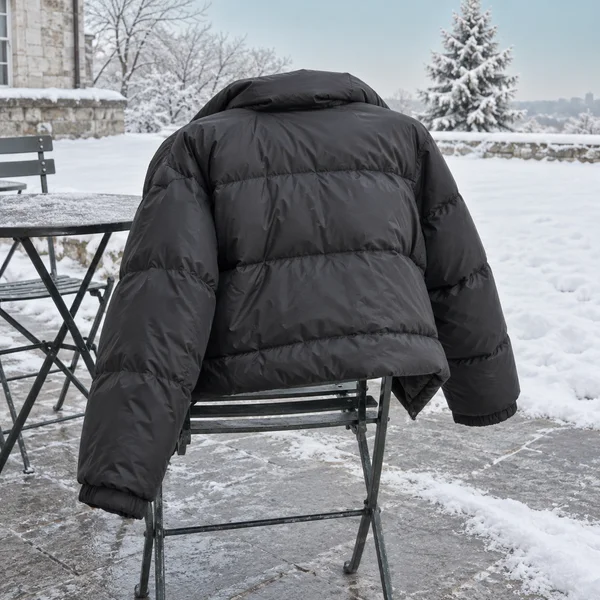 A dark charcoal puffer jacket hangs over a narrow metal café chair on a snow-dusted terrace, with a metal table, stone building, bare trees, and snow-covered…