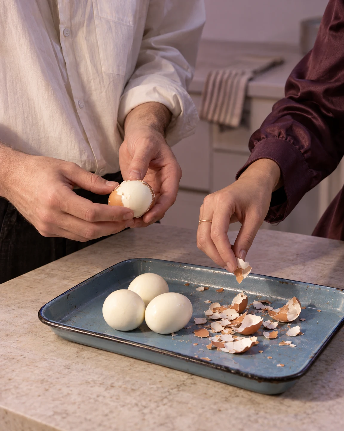 Two cropped adults peel hard-boiled eggs over a blue enamel tray on a kitchen worktop; one holds a partly peeled egg while the other drops a shell fragment…
