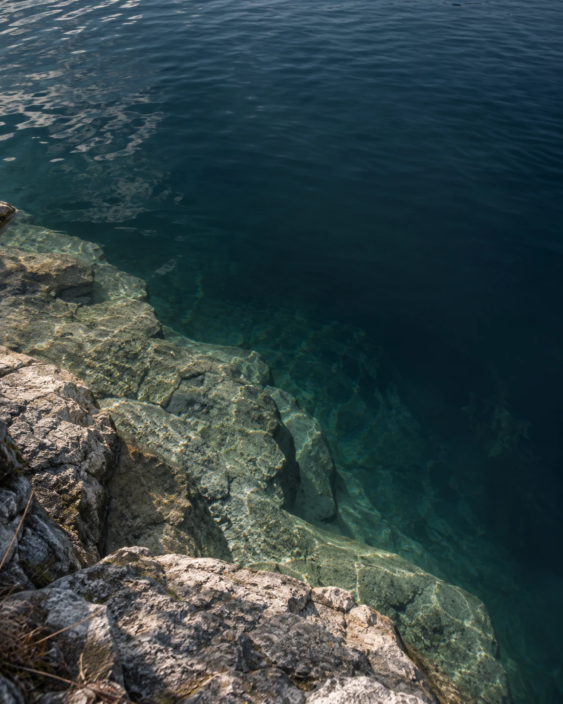 An oblique view down from a rocky bank onto a broad, pale stone shelf beneath clear lake water, beside a deep blue-green basin with surface ripples and…