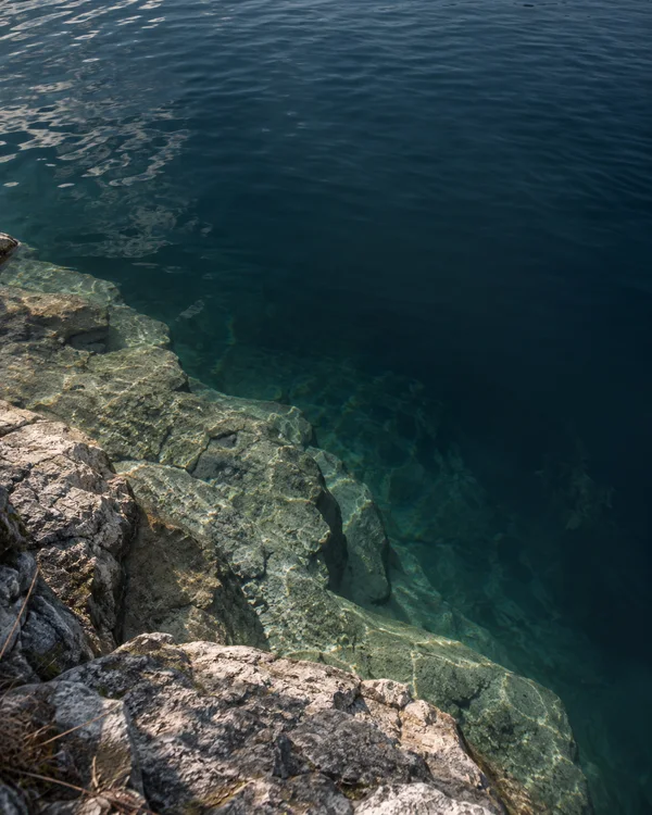 An oblique view down from a rocky bank onto a broad, pale stone shelf beneath clear lake water, beside a deep blue-green basin with surface ripples and…