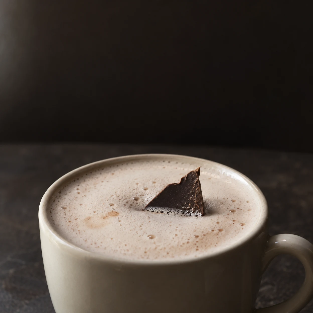 A large off-white ceramic cup filled with pale foamy hot chocolate sits on a dark café tabletop, with a jagged triangular piece of chocolate standing partly…