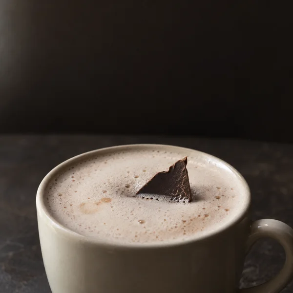 A large off-white ceramic cup filled with pale foamy hot chocolate sits on a dark café tabletop, with a jagged triangular piece of chocolate standing partly…