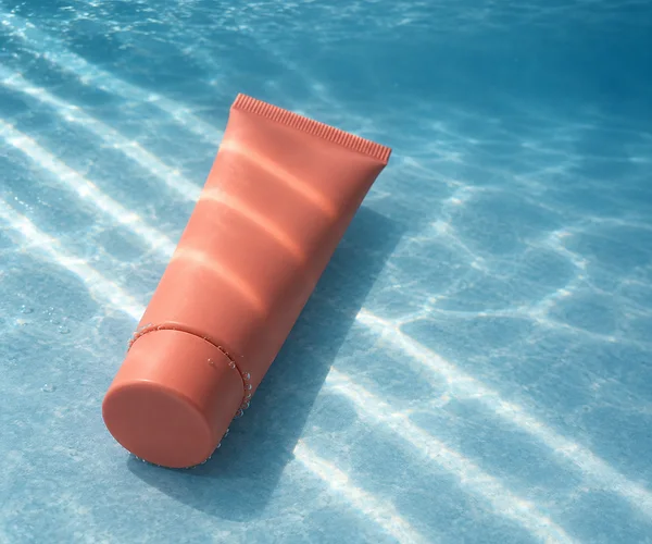 A blank coral squeeze tube lies diagonally on a pale blue underwater floor, with its cap in the lower left and sealed crimp toward the upper center; bright…