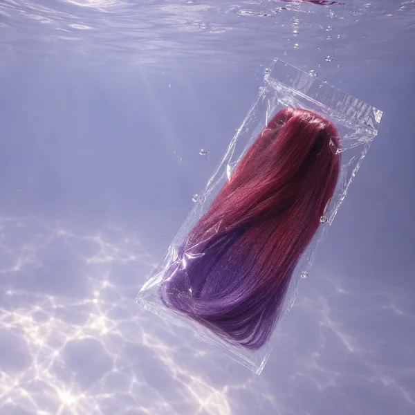 A transparent sealed pouch containing long berry-red and violet synthetic braiding fibers hangs diagonally underwater against pale lavender water, with bubbles…