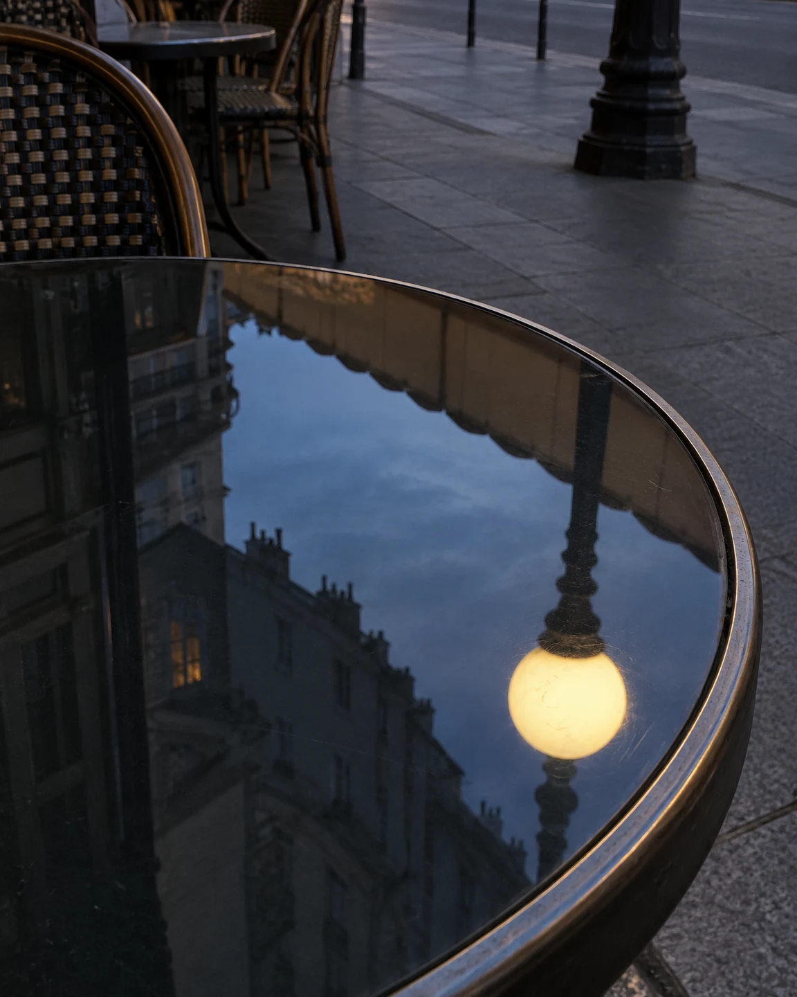 An empty glass-topped café table reflects a glowing globe streetlamp, blue dusk sky and surrounding buildings beside a paved street, with café chairs and a…