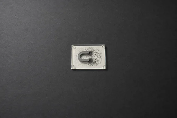 A small horseshoe magnet and scattered iron filings sit beneath a square glass plate with clear corner spacers, centered on a broad charcoal textured surface.