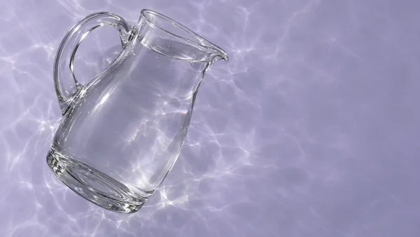 A clear glass serving pitcher lies diagonally across the left side of a pale lavender surface, with its curved handle and pouring lip visible.
